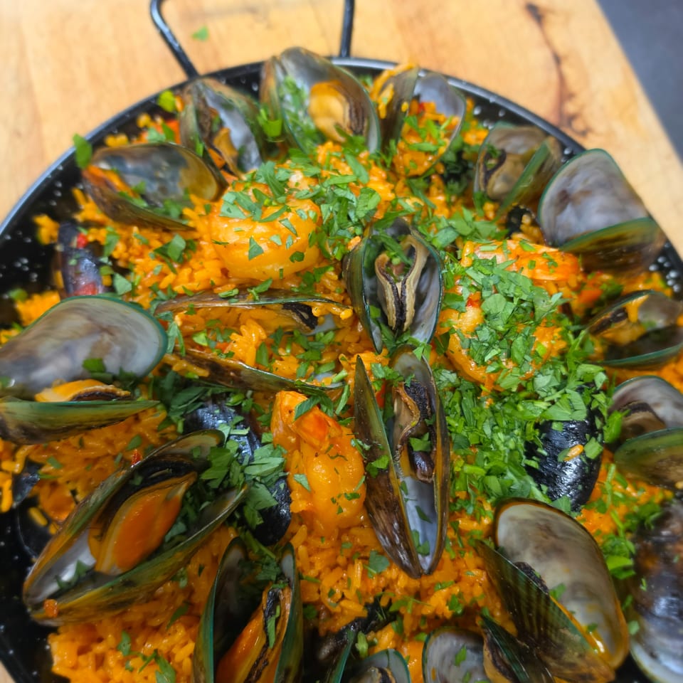 Black skillet filled with orange seafood rice, mussels, clams, shrimp, and chopped parsley on a wooden table.